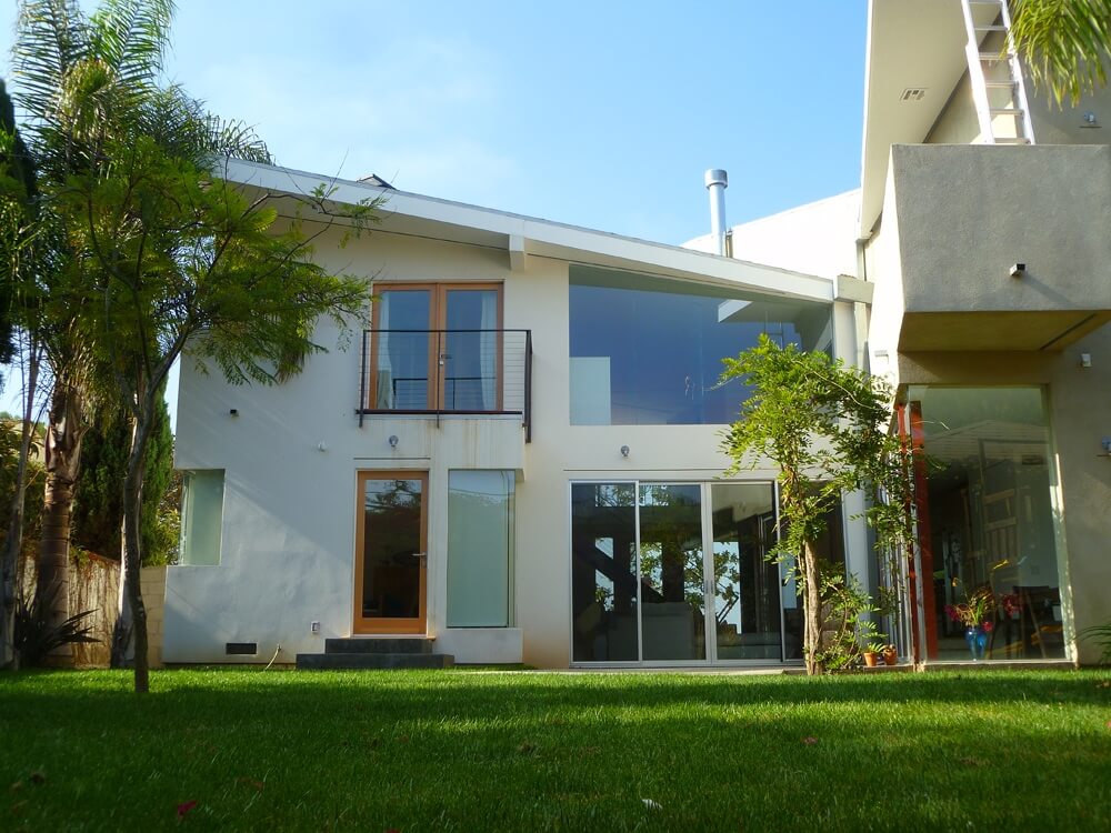 Backyard exterior of the Kerner Ho Residence with expansive glass, balcony, and modern white stucco forms surrounded by landscaped lawn and trees.