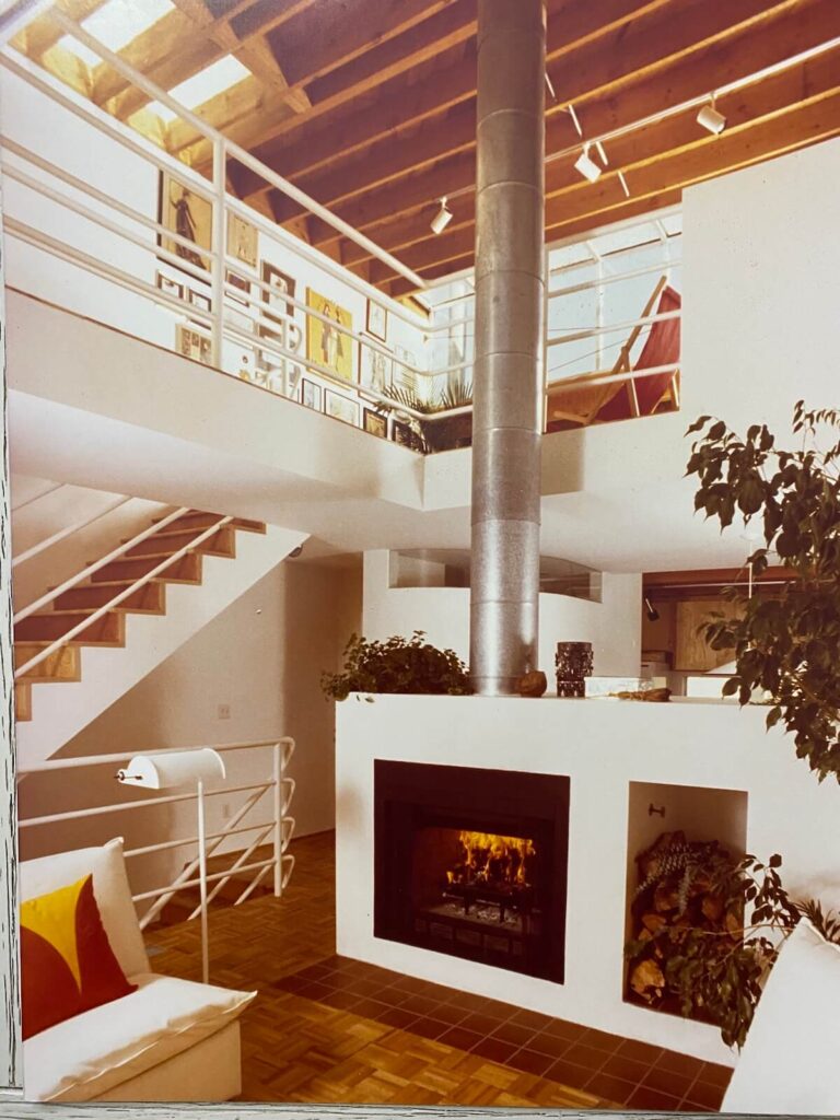 Double-height living room at the Pier Street Condos in Santa Monica with exposed wood ceiling, central fireplace, and wraparound mezzanine railing.