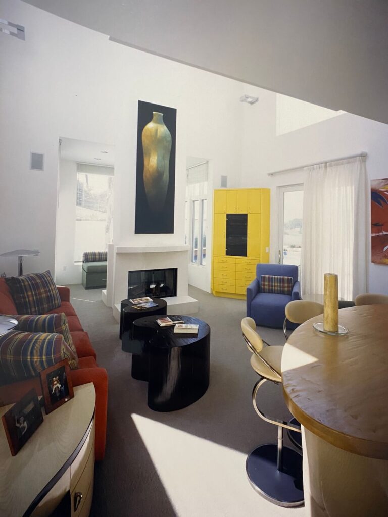 Living room inside the Shore Residence showcasing modern furnishings, a central fireplace, tall windows, and vibrant accent cabinetry.